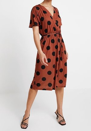 Vrouw draagt roestkleurige midi-jurk met zwarte polkadots, korte mouwen, V-hals en getailleerde taille, gecombineerd met zwarte sandalen met bandjes en hak.
