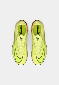 Jasné žlté futbalové kopačky s textúrovaným zvrškom, čiernym logom Nike a živými oranžovými akcentmi na päte. Šnurovací dizajn.
