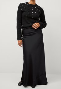 Schwarzer Strickpullover mit perlenartigen Akzenten auf der Vorderseite, kombiniert mit einem eleganten schwarzen Satin-Maxikleid und schlichten schwarzen Schuhen.