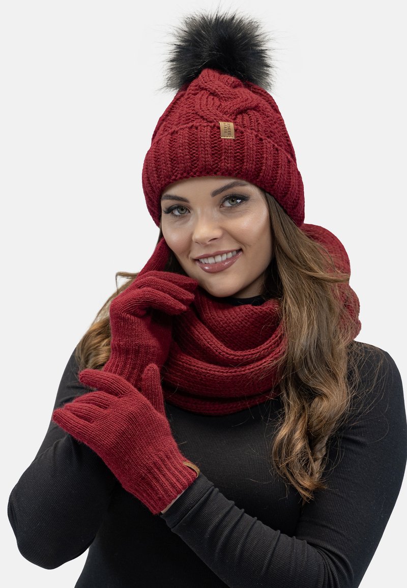 Rote Strickmütze mit einem schwarzen Fellpompon, passendem Schal und Handschuhen; strukturierter Stoff; enganliegendes Design.