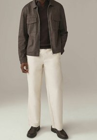 Bruine gestructureerde jas met ritssluiting en twee borstzakken, gecombineerd met een donkere trui en crèmekleurige wijde broekspijpen. Bruine schoenen maken de outfit compleet.