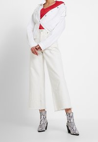 Giacca di jeans bianca cropped con dettagli in metallo argentato, top rosso con scollo a V, pantaloni bianchi a gamba larga e stivaletti con stampa serpente e tacco blocco.