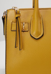 Gros plan d’un sac à main en cuir jaune moutarde texturé avec fermeture éclair dorée, pompon et détail de poignée cousue.