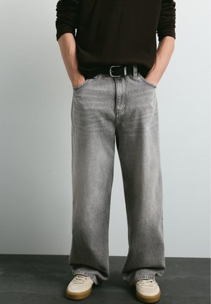 Une personne pose avec un jean gris à jambes larges, une ceinture noire et des baskets crème, les mains dans les poches.