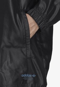 Veste noire et élégante avec des poignets élastiques, une poche latérale et une finition brillante. Présente le logo Adidas en bleu près de l'ourlet.