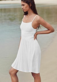 Robe d'été blanche avec un corsage smocké ajusté et des bretelles fines, s'évasant en une jupe légère et fluide. Texture lisse, design au genou.