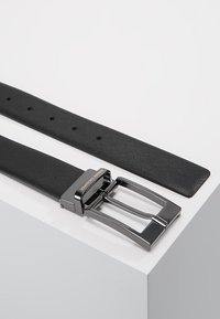 Ceinture en cuir noir avec une finition texturée, boucle en métal argenté avec un design à pointes et un logo gravé. Présentée sur une surface blanche.