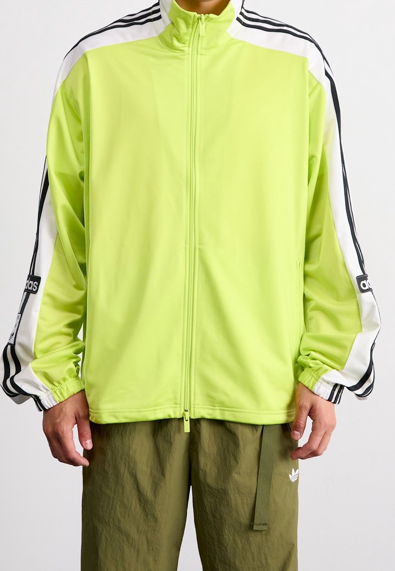 Neon green zippered jacket with white and black striped accents, elastic cuffs, and smooth texture. Features brand logo on sleeve.