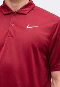 Άντρας που φοράει κόκκινο polo με κοντά μανίκια και δύο κουμπιά, με λευκό λογότυπο Nike στο στήθος.