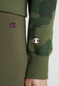 Felpa verde con polsini a coste e maniche con motivo camouflage. Presenta il logo ricamato Champion in blu e bianco sulla manica e in vita.