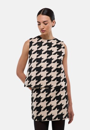 Femme portant un haut sans manches et une jupe avec un grand motif pied-de-poule noir et beige, debout devant un fond clair uni.