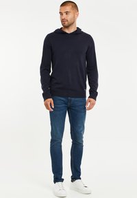 Navy-Strickhoodie mit langen Ärmeln und vorderer Tasche, kombiniert mit schlanken blauen Jeans und weißen Sneakers. Glatte Textur, lässiges Design.