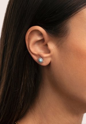 Boucles d'oreilles en stud turquoise avec un motif en éclat de soleil, montées en métal argenté. Présentent une surface lisse et de petits accents transparents autour du bord.