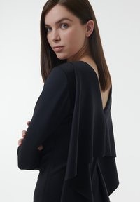 Zwarte jurk met lange mouwen, voorzien van een open rug en een vloeiende gelaagdheid bij de schouders. Soepele stoftextuur.