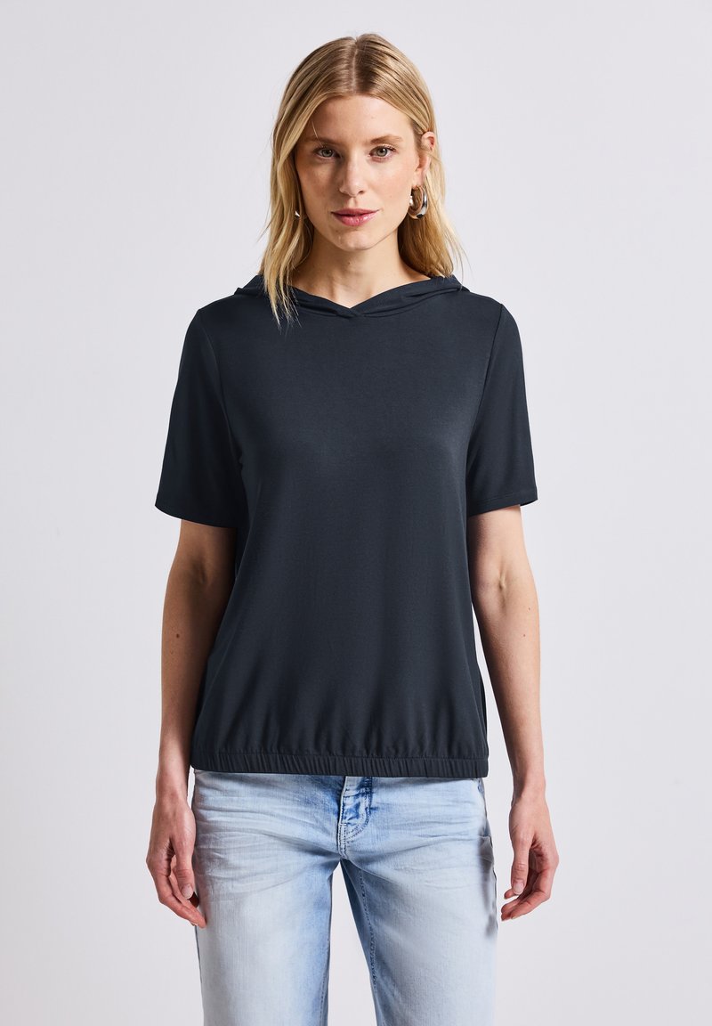 Top negro de manga corta con capucha y dobladillo elástico; tejido suave y ligero; modelo lleva puestos unos jeans de color azul claro.