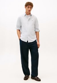 Camicia azzurra chiara a maniche lunghe con maniche arrotolate, pantaloni blu navy e mocassini marroni. Vestibilità casual, tessuto texturizzato, dettagli con piccolo logo.