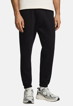 Mann trägt schwarze Jogginghose und weiße Sportschuhe mit grauen und schwarzen Details, steht vor einem schlichten hellen Hintergrund.
