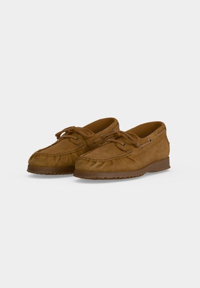 Paire de mocassins en daim marron avec lacets et semelles en caoutchouc, montrée en angle sur un fond clair uni.
