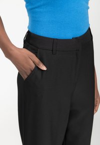 Zwarte op maat gemaakte broek met zijzakken, zachte stof en een goed passende tailleband. Een blauwe top is gedeeltelijk zichtbaar boven de tailleband.