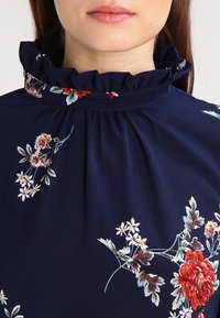 Žena v námořnickém modrém blůzce s volánkovým vysokým límcem a červeným, bílým a zeleným květinovým vzorem, s ústy zavřenými a neutrálním výrazem.