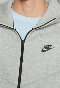 Harmaa vetoketjullinen huppari, pehmeästä kankaasta valmistettu. Varustettu mustalla vetoketjulla ja Nike-logolla, vaaleansininen t-paita näkyy hupun alla.