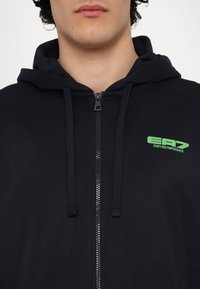 Navy zip-up hoodie made of soft fabric, featuring a green logo on the left chest, with drawstrings and a standard hood design.
