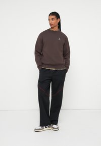 Hombre con trenzas largas que lleva puesta una sudadera marrón, una camiseta de camuflaje debajo, pantalones negros con costuras rojas y zapatillas grises y blancas, de pie.