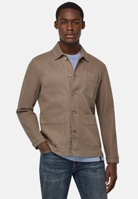 Giacca di tela marrone con bottone frontale, colletto a punta, due tasche frontali e una tasca sul petto. Indossata sopra una t-shirt azzurro chiaro.