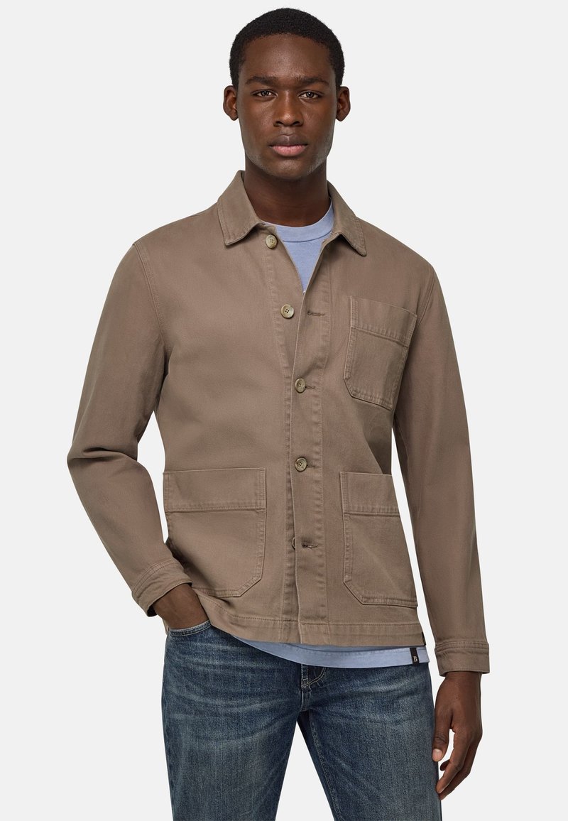 Giacca di tela marrone con bottone frontale, colletto a punta, due tasche frontali e una tasca sul petto. Indossata sopra una t-shirt azzurro chiaro.