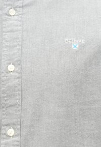 Camisa abotoada cinza-claro com o logótipo da Barbour bordado em branco e azul no peito, perto dos botões.