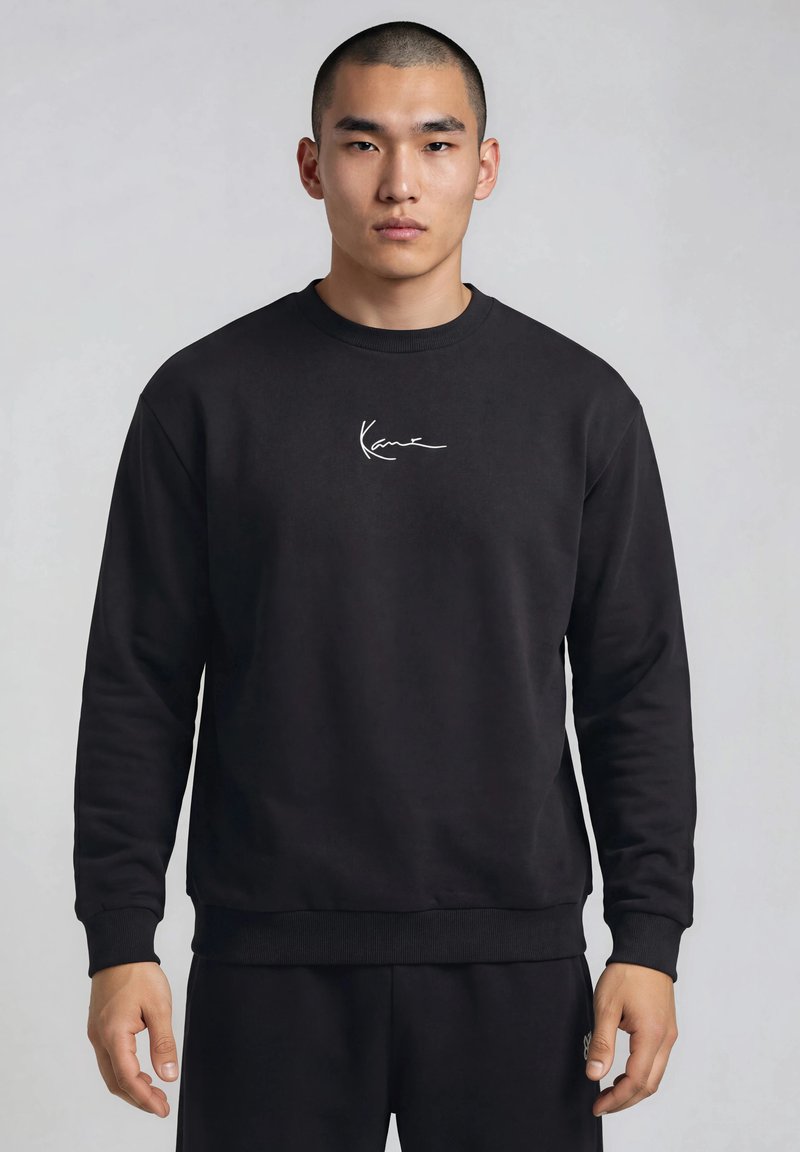 Homme aux cheveux courts portant un sweat-shirt noir avec un petit logo blanc en écriture centré sur la poitrine, debout devant un fond gris uni.