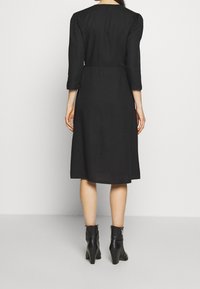 Schwarzes knielanges Kleid mit Dreiviertel-Ärmeln, taillierter Passform und glatter Textur. Kombiniert mit schwarzen, hohen Stiefeletten.
