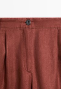 Pantalon en tissu couleur rouille avec un bouton noir et des coutures visibles à la taille et sur la braguette avant.