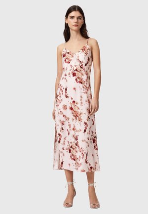 Kvinde iført en ærmeløs pink midi-kjole med blomsterprint, spaghetti-stropper og hvide ankelbindte hæle, stående mod en hvid baggrund.