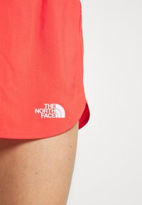 Röda sportshorts i texturerat tyg, med en vit logotyp från The North Face nära fållen. Designen inkluderar en rundad kant.