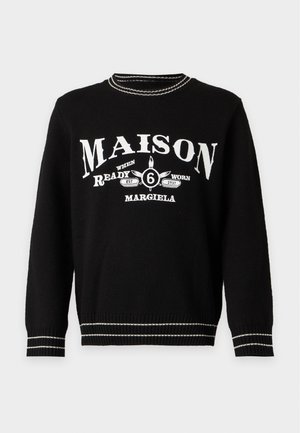 Junger Mann trägt schwarzen Pullover mit weißem "Maison Margiela"-Text, schwarze Hose und dunkelrote Stiefel mit roten Schnürsenkeln, steht vor weißem Hintergrund.