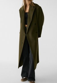 Olijfgroen oversized wollen jas met brede revers, dubbelknopen en een middellange stijl, gedragen over een zwart cropped top en jeans.