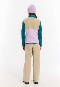 Fleece jack in teal, beige en lavendel; ritsontwerp met hoge kraag. Beigepantalon en paarse gebreide beanie. Zwarte schoenen.