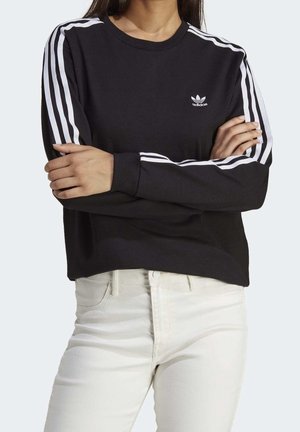 Kobieta ubrana w czarną bluzę Adidas z białymi paskami na rękawach i logo na piersi, w połączeniu z białymi spodniami, ramiona skrzyżowane.