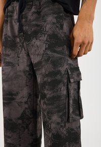 Close-up of a person's hand near black and gray camo-patterned cargo pants with a flap pocket on the side.