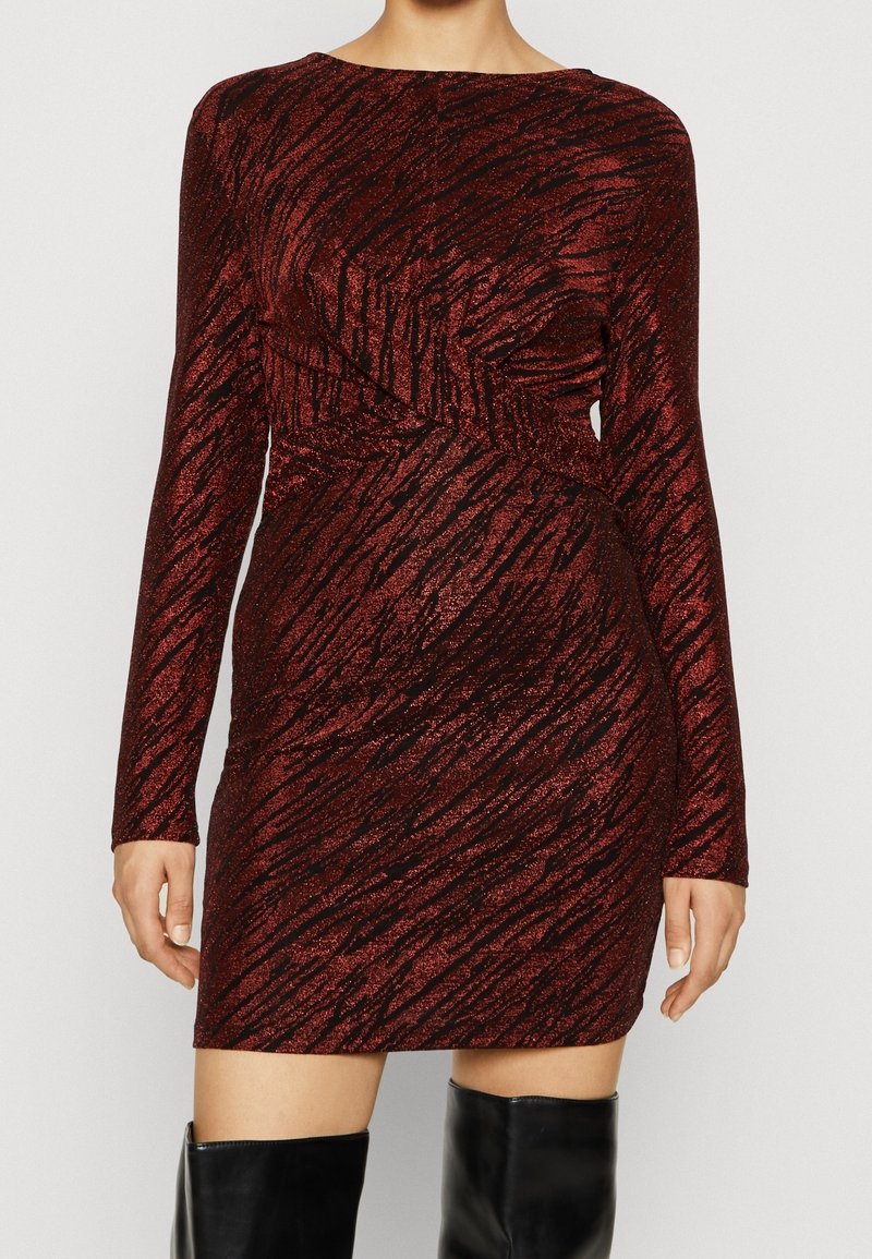 Femme portant une mini-robe à manches longues, ajustée, scintillante avec un motif rouge foncé et noir, col bateau, et des bottes noires montantes jusqu'aux genoux.