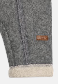 Tela de forro polar gris con costura visible y forro de sherpa cream doblado, que presenta una pequeña etiqueta de marca marrón cosida.