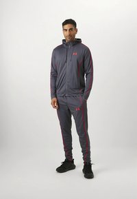 Grijze sportieve trainingspak gemaakt van lichtgewicht materiaal, met rode accenten en een hoodie met rits. Inclusief joggerbroek met zijstrepen.