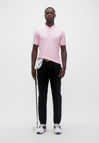 Άνδρας με ροζ πόλο, μαύρο παντελόνι και παπούτσια γκολφ, κρατώντας γκολφ ρόπαλο με άσπρο γάντι περασμένο μέσα στο παντελόνι, σε λευκό φόντο