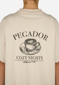 Bēšas kokvilnas krekls ar drukātu kafijas krūzes grafiku ar sirds formas dizainu, virsū uzraksts "Pegador Cozy Nights."