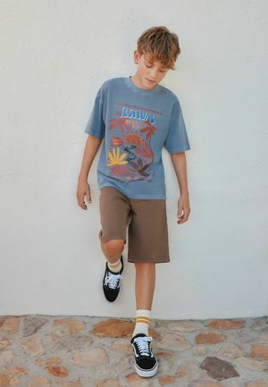 Băiat cu tricou albastru cu grafică și pantaloni scurți maro ridică un picior stând pe o pavăză de piatră în fața unui perete alb.