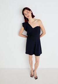 Vestido azul-marinho, de um ombro só, em tecido suave com um detalhe de renda na manga, design ajustado na cintura e saia em formato de campânula, com comprimento acima do joelho.