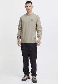 Sudadera de color beige claro con cuello redondo y un pequeño parche de logo negro, combinada con pantalones cargo negros y zapatos de senderismo grises.