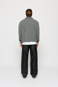 Person som står med ryggen til, iført en grå strikkegenser, hvit skjorte, svarte skinnoble bukser og sneakers mot en ensfarget bakgrunn.