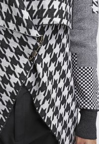Černobílý pletený kardigan se vzorem houndstooth, ozdobnými knoflíky a kontrastními kostkovanými rukávy.